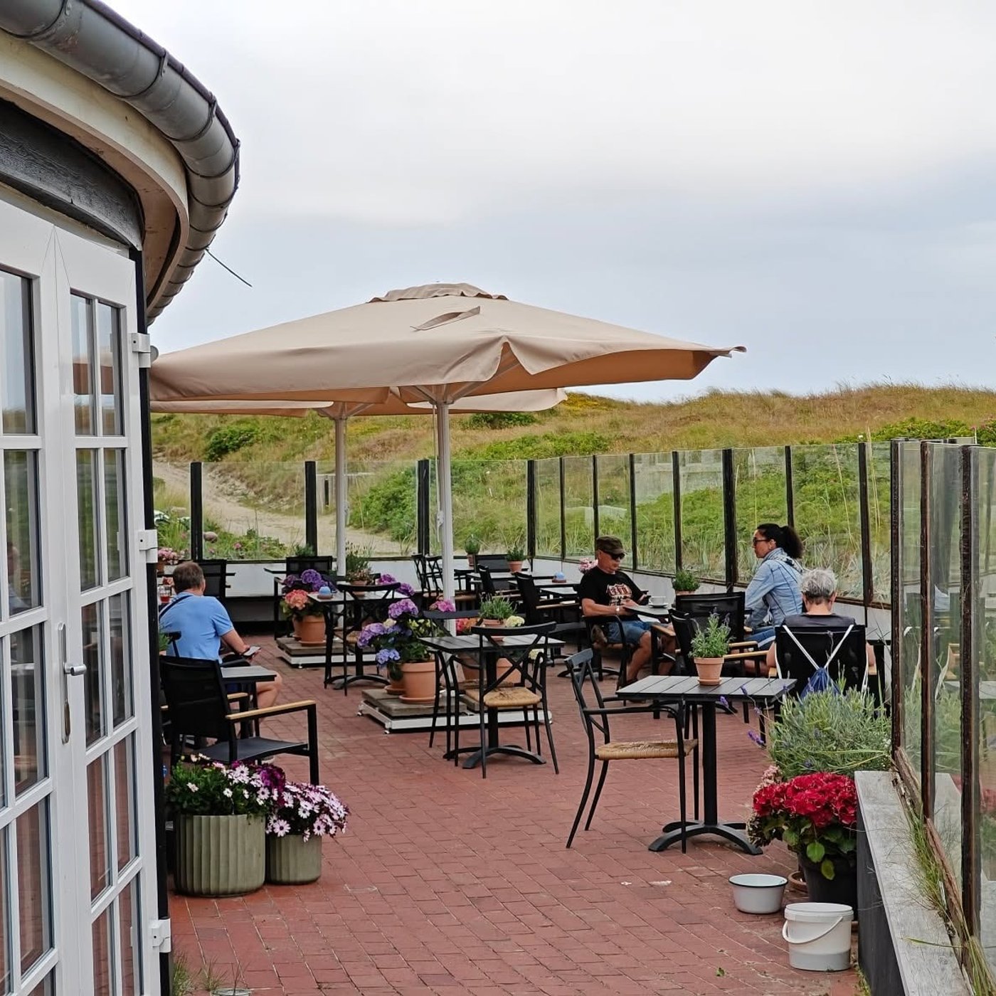 Udendørs caféterrasse med borde, parasoller og gæster tæt ved klitterne